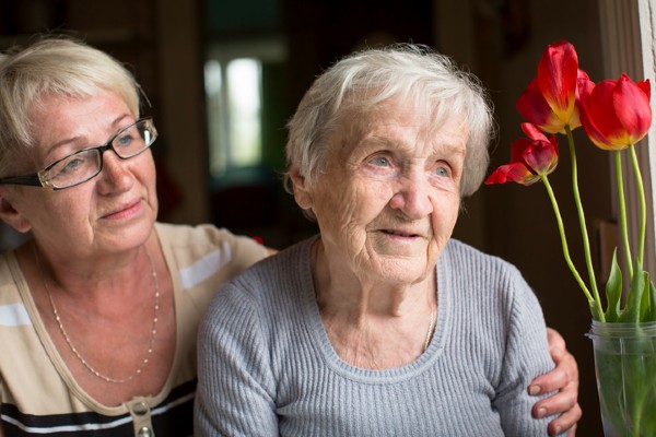 A woman puts her arm around her older mother, who looks out a window.