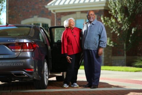 A senior couple stands by a car in front of a senior living community.