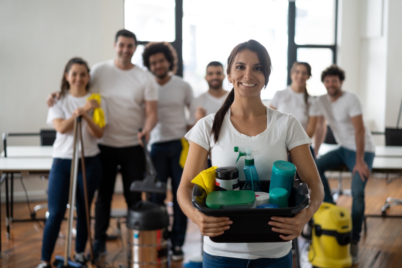 Beautiful cleaning woman with her team at an office holding a bucket with cleaning products all smiling at camera