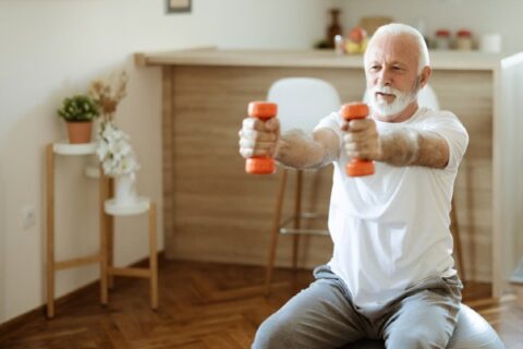 A senior man does resistance training with dumbbells in his home.