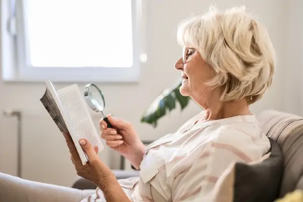 Senior Woman At Home Reading Book Using Magnifying Glass