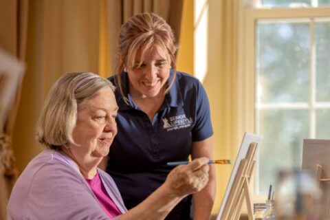 A senior paints while a caregiver watches.
