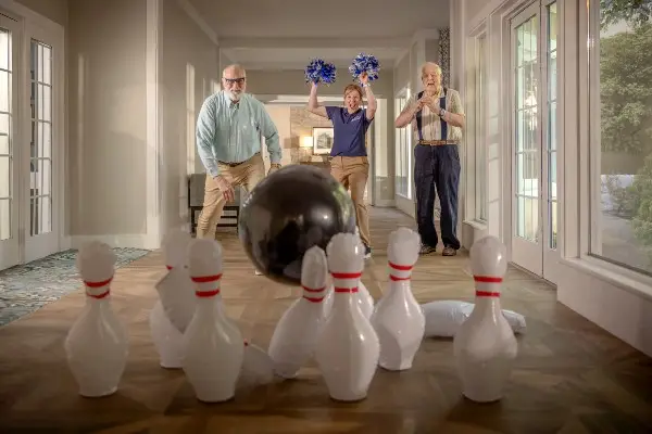 Two senior men play hall bowling with a caregiver at their Senior Lifestyle community.