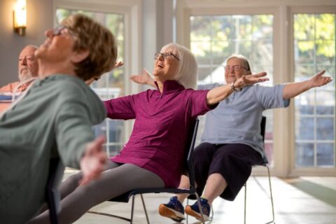 A group of smiling seniors participate in a chair yoga exercise class.