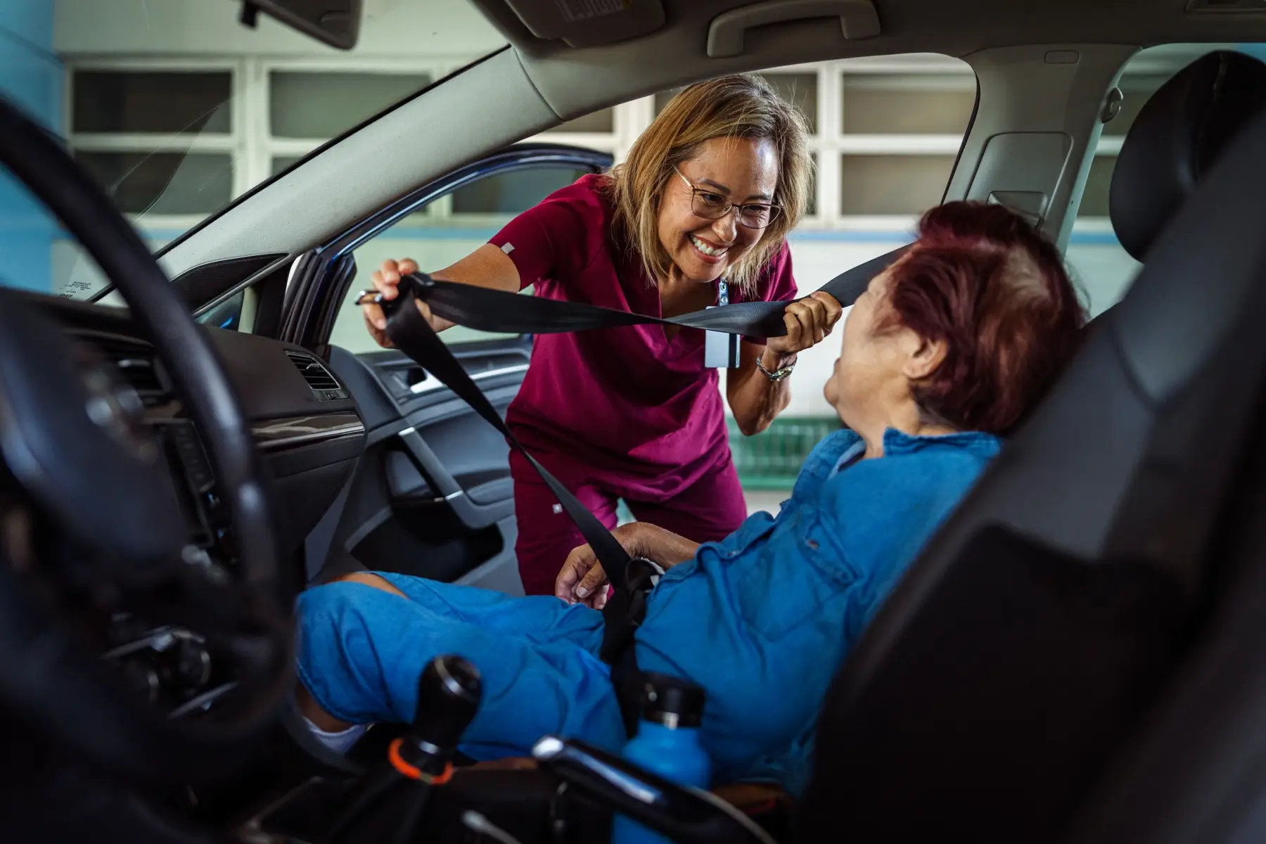 Home caregiver helping elderly patient get seated in passenger seat of parked car A kind occupational therapist of Hawaiian and Finnish descent smiles as she helps a female geriatric patient get seated and buckled into the passenger seat of a car.