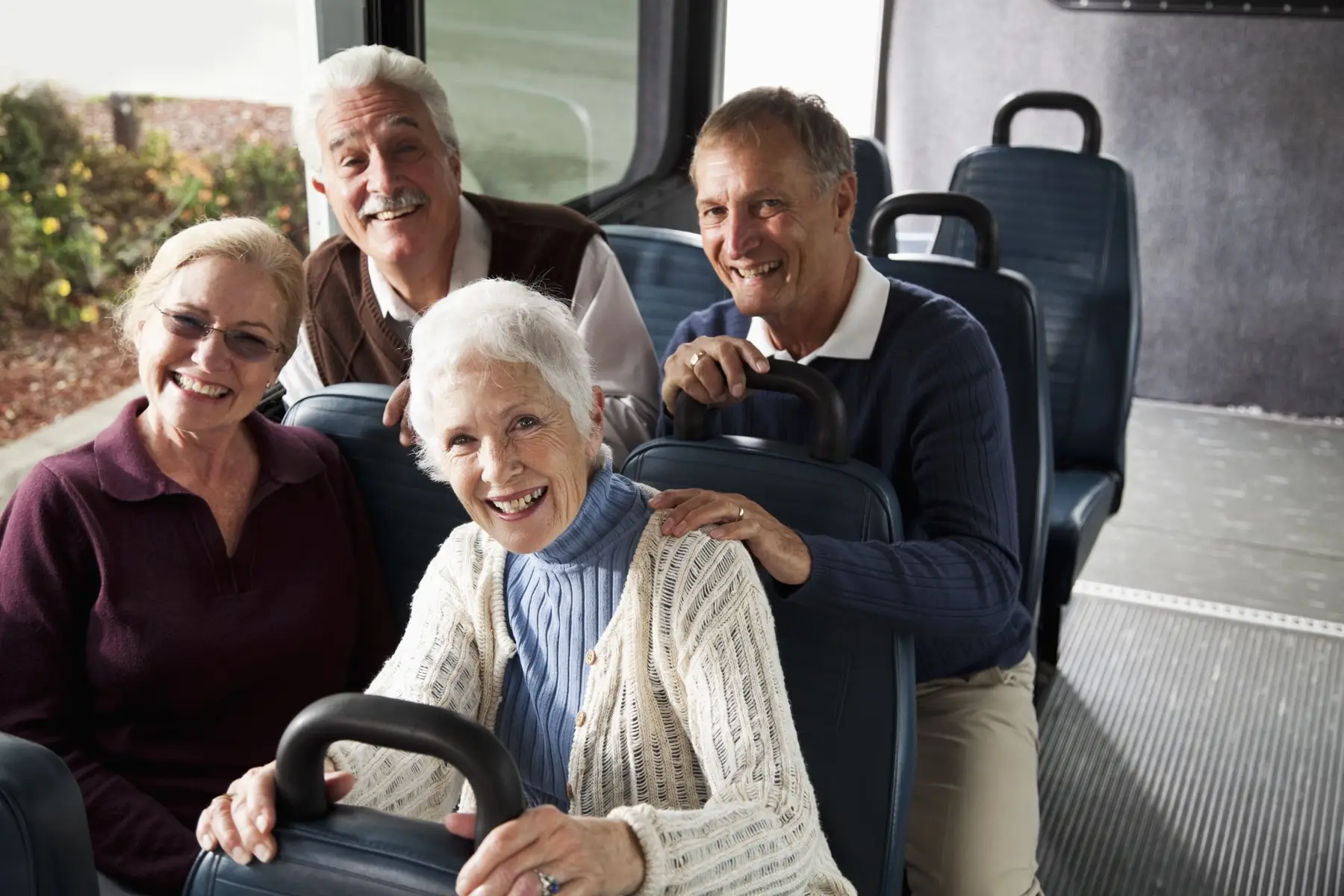 Seniors in shuttle bus A group of four senior citizens riding in a van or shuttle bus, smiling at the camera. The focus is on the woman with white hair in the foreground, wearing a white sweater. Two women are sitting in front and two men are sitting directly behind them. They are friends riding together, two couples.